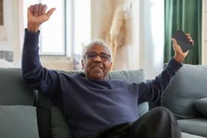 Senior man joyfully sitting on a couch holding a smartphone indoors.